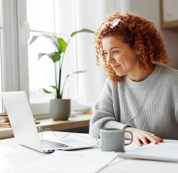 Cute university student with curly red hair doing homework sitting at kitchen table with coffee cup and wired earphones next to big window, watching online tutorial on laptop and writing in copybook