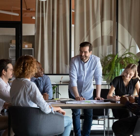 Happy male business team leader talking to employees at corporate meeting, discussing work project, sharing jokes, laughing. Positive mentor training diverse group of interns, having fun