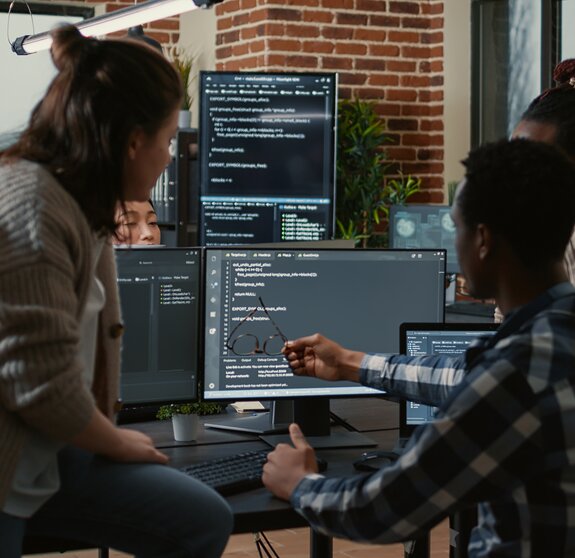 Programer sitting on desk discussing with mixed team of software developers about artificial intelligence innovation. Programmers doing teamwork looking at running machine learning algorithms.