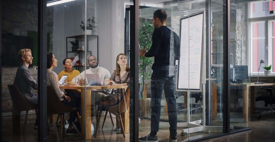 Project Manager Makes a Presentation for a Young Diverse Creative Team in Meeting Room in an Agency. Colleagues Sit Behind Conference Table and Discuss Business Development, User Interface and Design. 