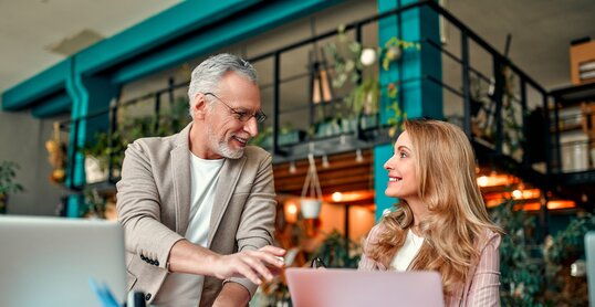 Frau und Mann sitzen gemeinsam an Schreibtisch und unterhalten sich