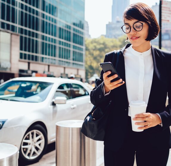 Concentrated female entrepreneur using application for calling taxi getting to work with morning coffee to go, prosperous businesswoman checking notification on smartphone strolling with takeaway.