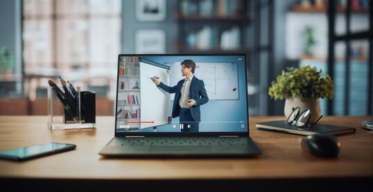 Shot of a Laptop Computer Showing Online Lecture with Portrait of a Cute Male Teacher Explaining Math Formulas. It is Standing on the Wooden Desk in Stylish Modern Home Office Studio During Day.