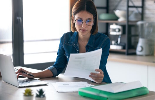 Young business woman working with computer while consulting some invoices and documents in the kitchen at home.