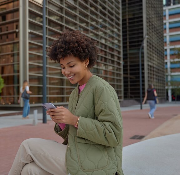 Sideways shot of beautiful woman with Afro hair holds smartphone reads received sms message has cheerful expression sits in downtown against cityscrapers dials number of taxi service. Technology