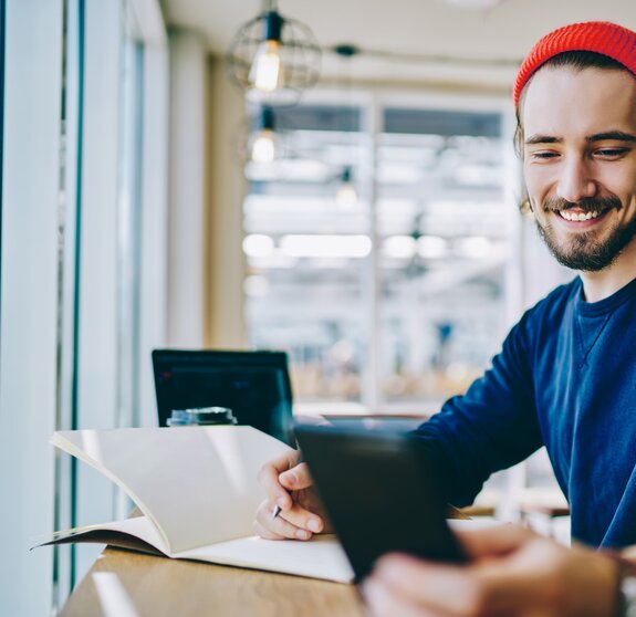 Caucasian male student connected to 4g for browsing internet on smartphone device during studying with textbook for education, successful hipster guy checking notification on cellular phone