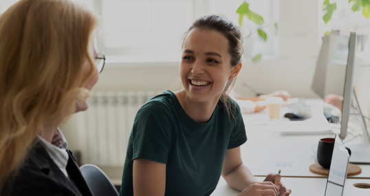 Pleasant workday. Two women colleagues employees of diverse age sharing points of view at workplace in office, happy smiling millennial female trainee discussing business ideas with adult aged trainer