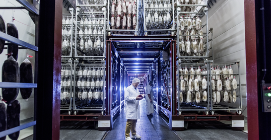 Foto einer Fabrikhalle der Bell Food Group mit zwei Mitarbeitern in Schutzkleidung und Waren die von Regalen hängen