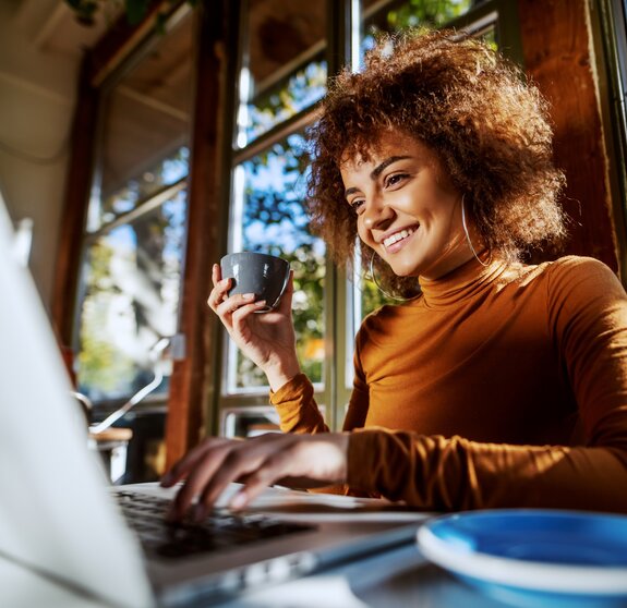 Charming young mixed race female freelancer with curly hair sitting in cafe, typing on laptop and holding cup of fresh coffee.